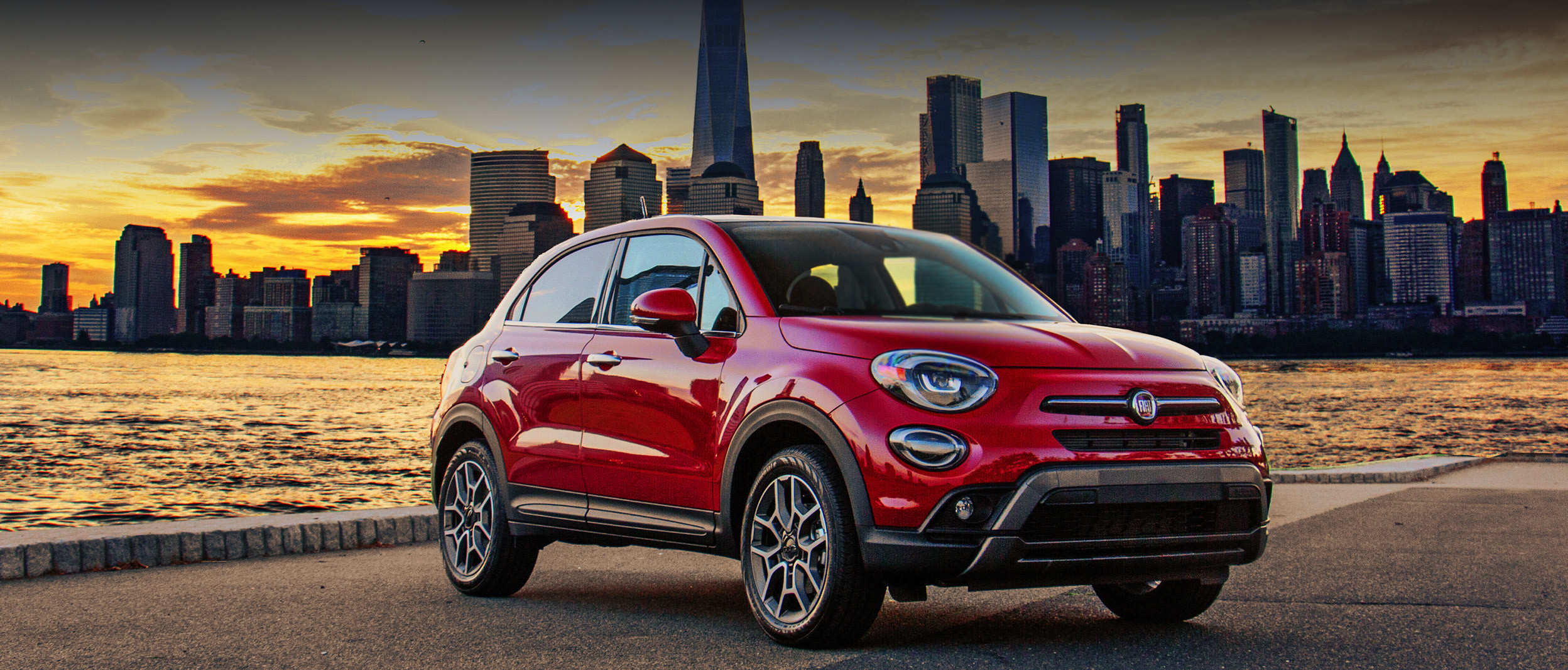 Une FIAT 500X 2020 rouge garée à l’extérieur près d’un lac devant un paysage urbain