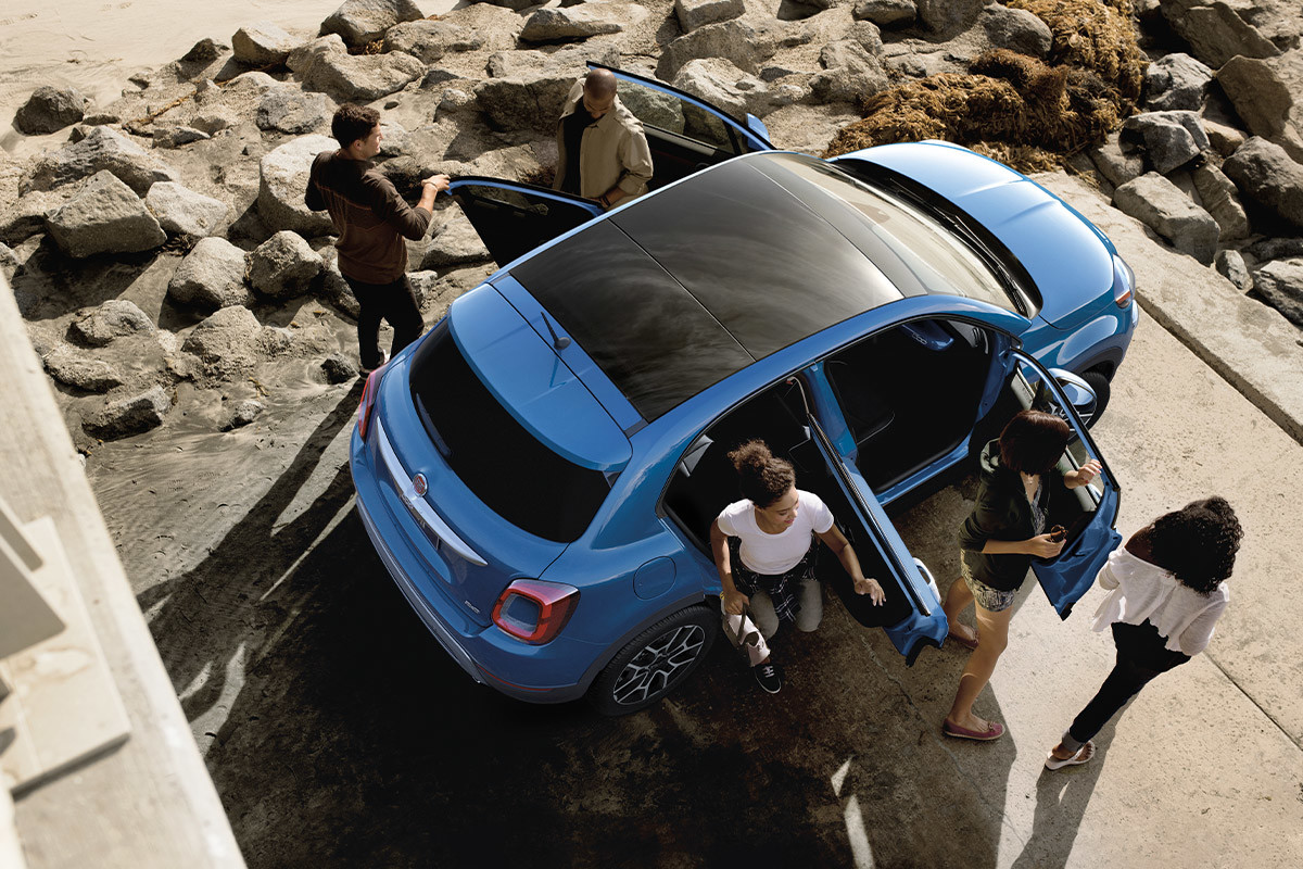 Vue du dessus d’une FIAT 500X bleue avec des personnes sortant du véhicule à la plage.