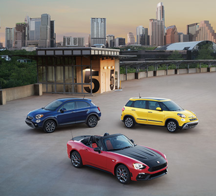 Red FIAT 124 Spider, yellow FIAT 500L, and blue FIAT 500X parked in a large empty lot with high rise buildings in the background.