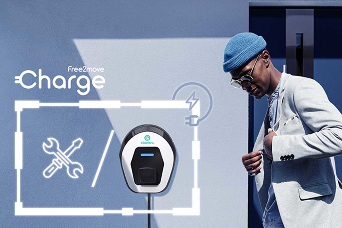 A man in a blue beanie and sunglasses walks past a white Free2move Charge EV charger mounted on a wall.