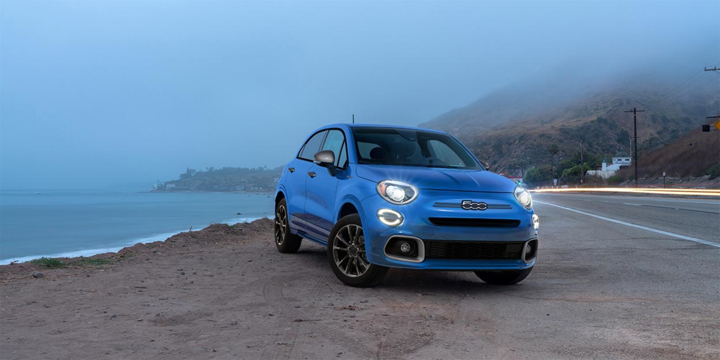 Vue de face d'une Fiat 2023 bleue garée sur un terrain sablonneux avec une vue sur l'océan et la chaussée montrée derrière.
