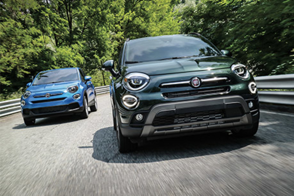 Une FIAT 500X verte et une autre bleue roulant sur une route pavée à travers des arbres verts.