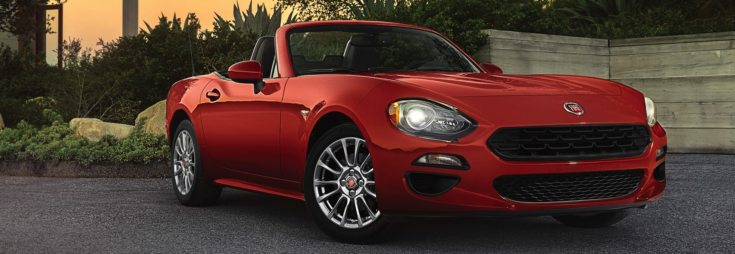 FIAT 124 Spider 2020 rouge stationnée dans une entrée en gravier, au coucher du soleil.