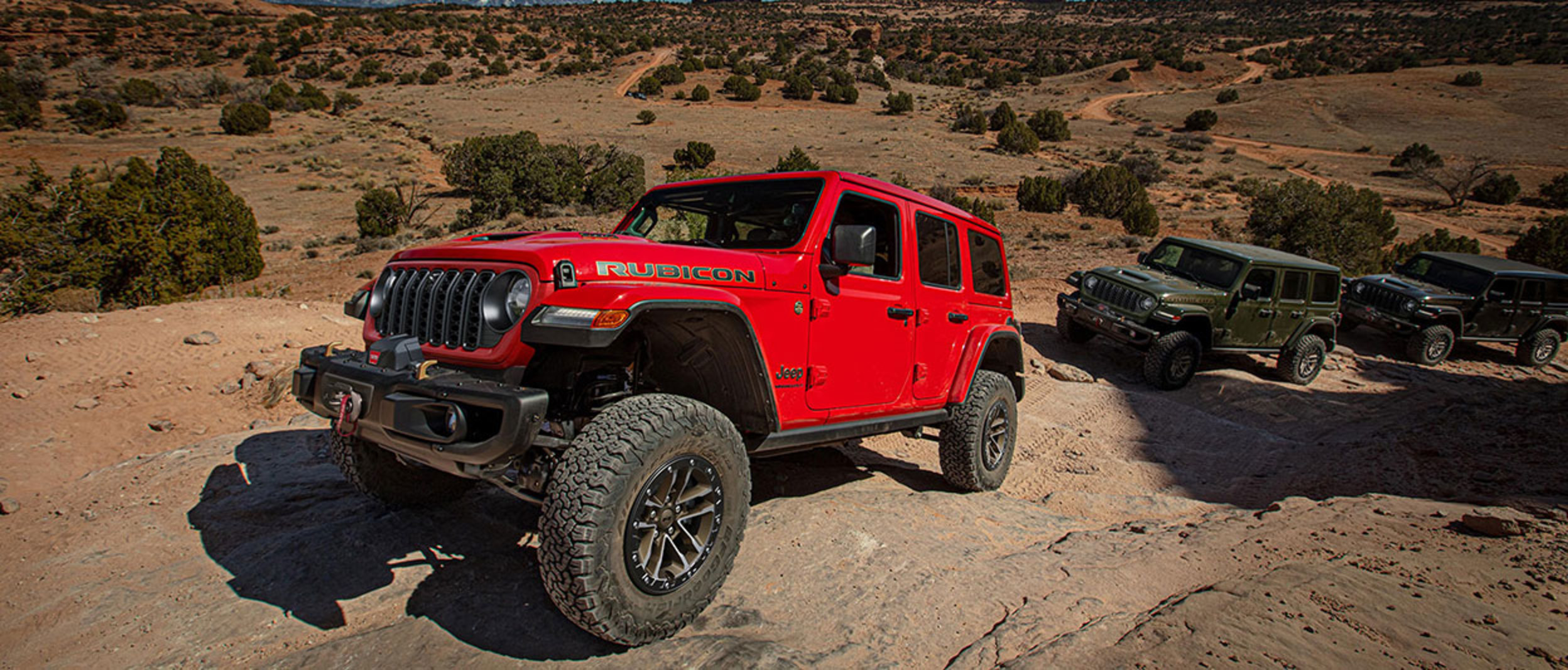 Angled view of a red Jeep Rubicon shown driving uphill on rocky terrain with two other Jeep models following behind.
