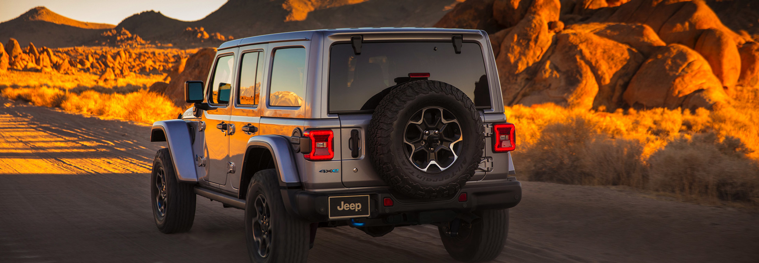 Vue arrière d'une Jeep Wrangler 4xe gris roulant sur un chemin de terre à travers un désert rocheux.