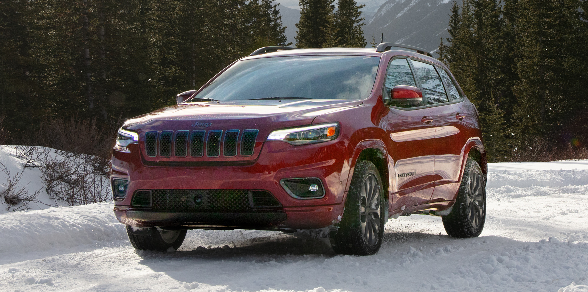 Red Jeep Cherokee on a snowy trail, against a backdrop of grey skies, trees and mountains.