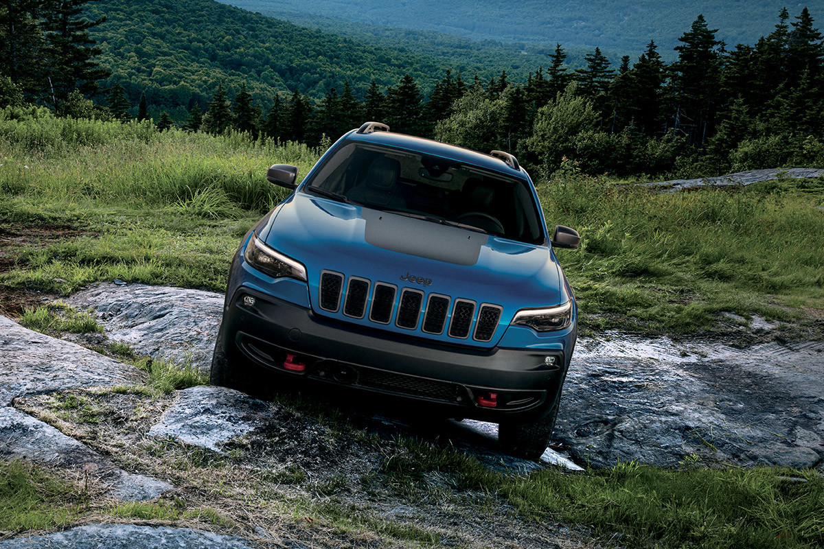 Front view of a 2022 Jeep Cherokee SUV driving on an off road trail with hills in the backdrop.