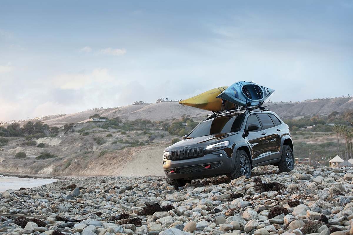 DESIGNED TO ELEVATE <span class="unbr">EVERY JOURNEY</span> 2022 Jeep Cherokee parked on a rocky shore with two kayaks mounted on roof rack cross bars.