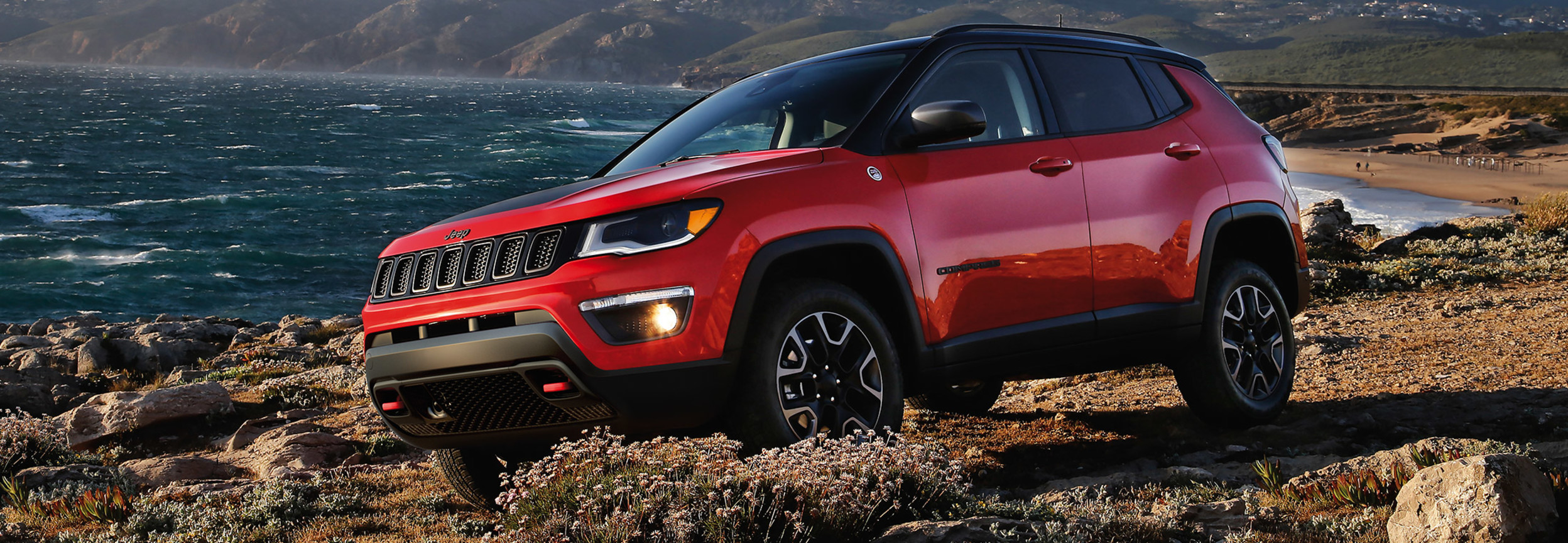 Vue latérale d’un Jeep Compass rouge stationné sur une falaise avec de l’eau en arrière-plan