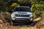 Silver Jeep Compass with black trim driving on a rocky trail with trees and shrubs in the background.