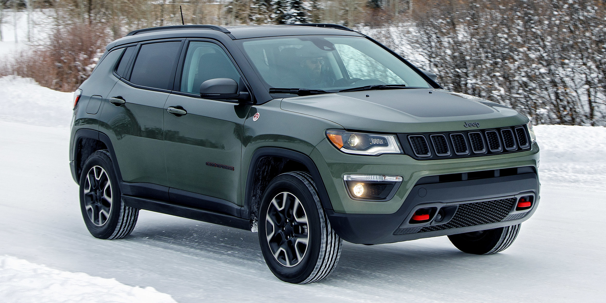 Compass Trailhawk vert olive et noir clair garé sur une route de campagne enneigée près d’un pont, des montagnes derrière.
