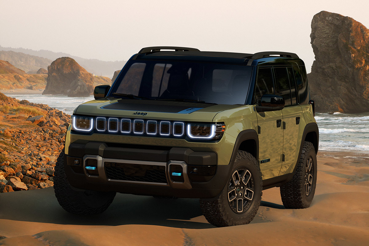 View of a green 2023 Jeep Recon model parked on top of sand terrain with water and rocks in the background.