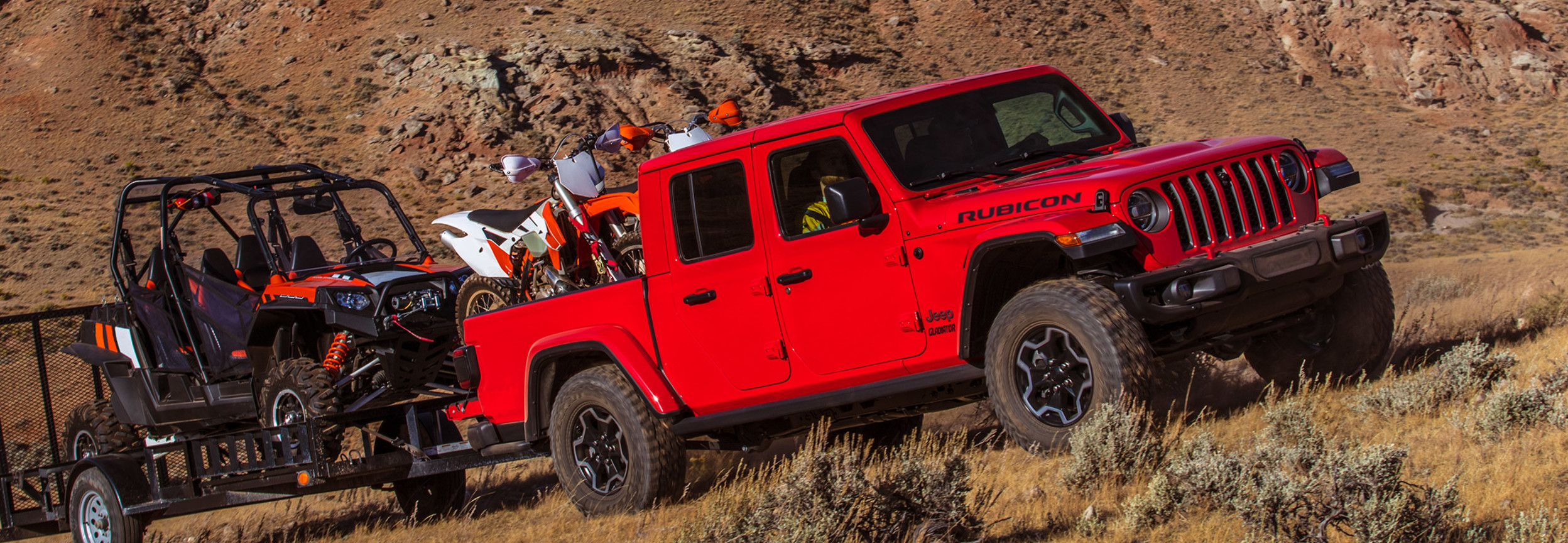 Vue d’un Jeep Gladiator rouge transportant deux motos et remorquant un véhicule de safari durant l’ascension d’une montagne