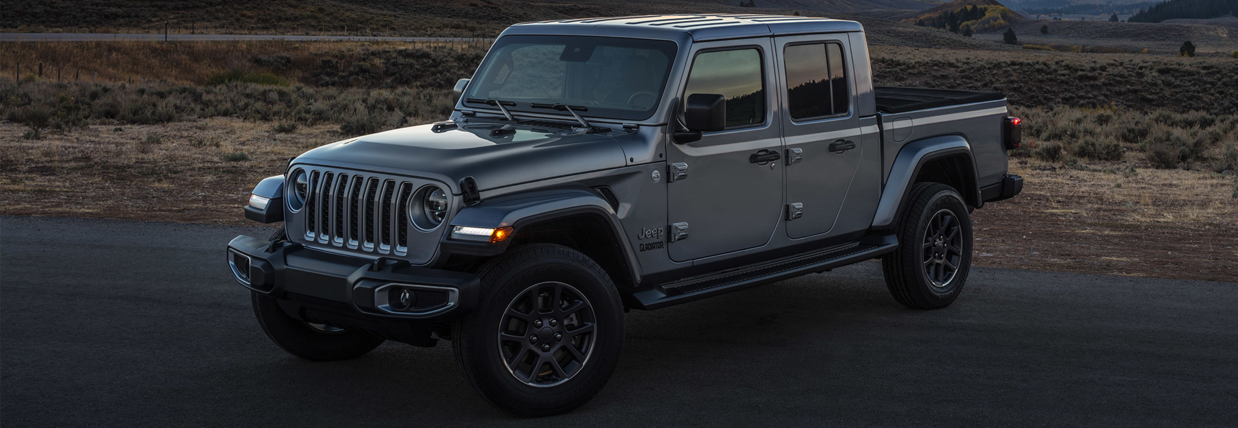 Vue latérale d’un Jeep Gladiator gris stationné sur une route avec des montagnes en arrière-plan