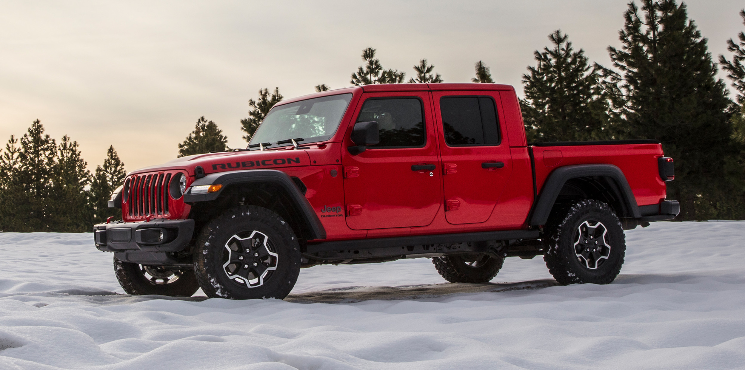 Red Jeep Gladiator Rubicon parked on an open, snow-covered landscape with a clear sunny sky and trees in the distance.