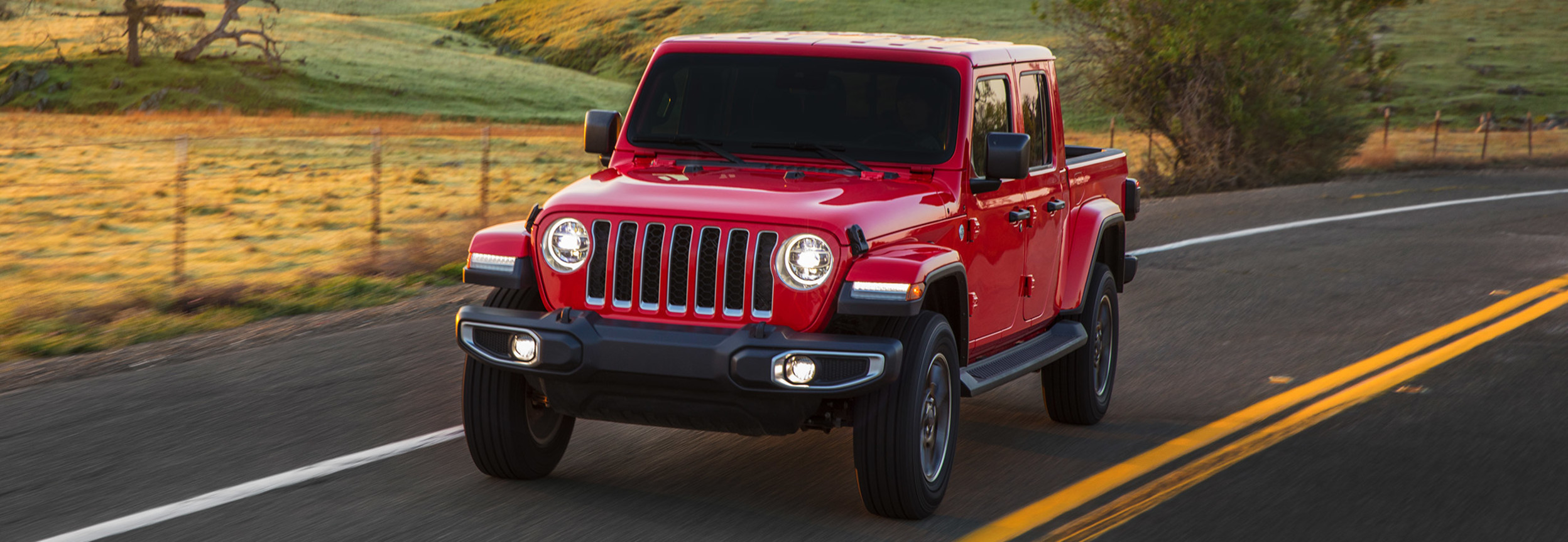 Red Jeep Gladiator driving down a road next to grassy hills and trees