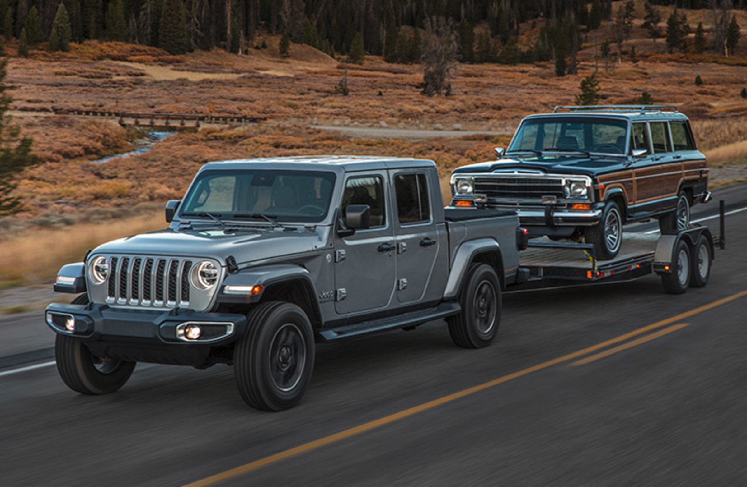 Vue avant d’un Jeep Gladiator 2021 gris pastenague tractant un Jeep Wagoneer rétro sur une remorque à l’arrière.