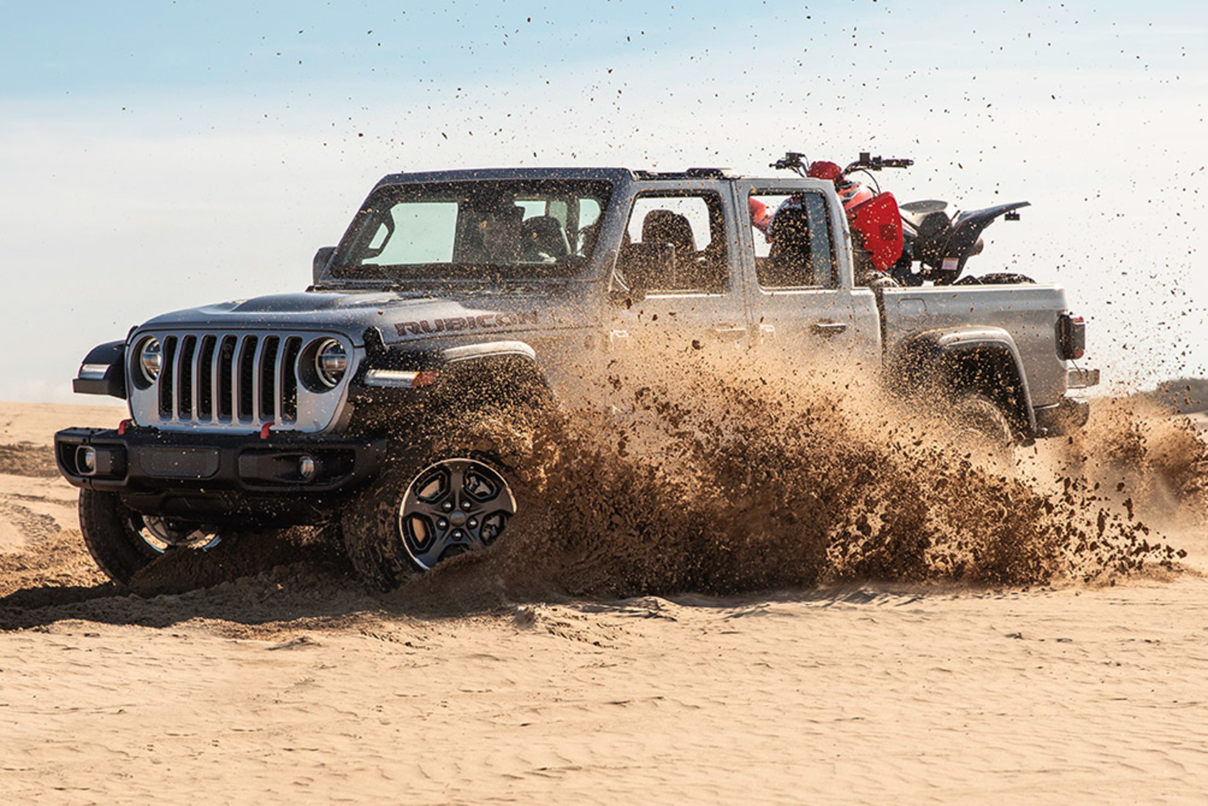 Vue latérale d’un Jeep Gladiator Mojave 2021 gris pastenague soulevant du sable en roulant dans le désert.