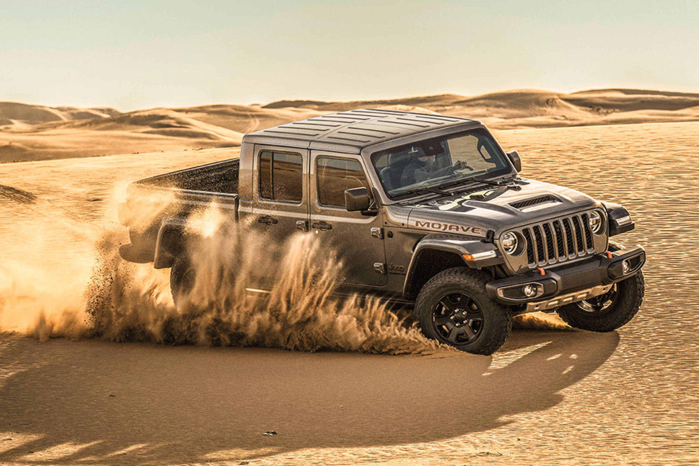 Vue latérale d’un Jeep Gladiator Mojave 2021 gris pastenague créant une crête de sable en tournant sur un terrain désertique.