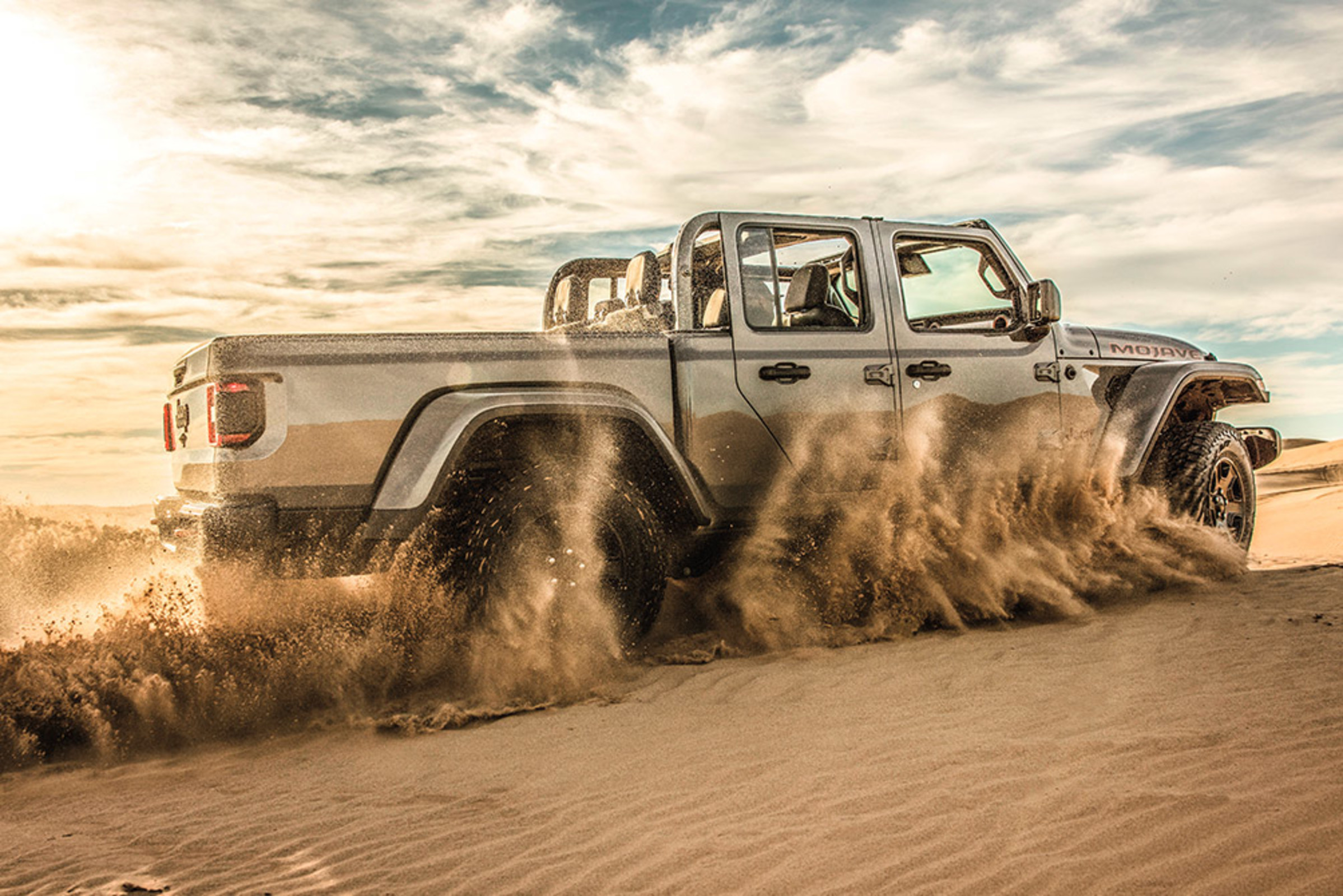 Vue latérale d’un Jeep Gladiator Mojave 2021 gris pastenague soulevant du sable en roulant dans le désert.