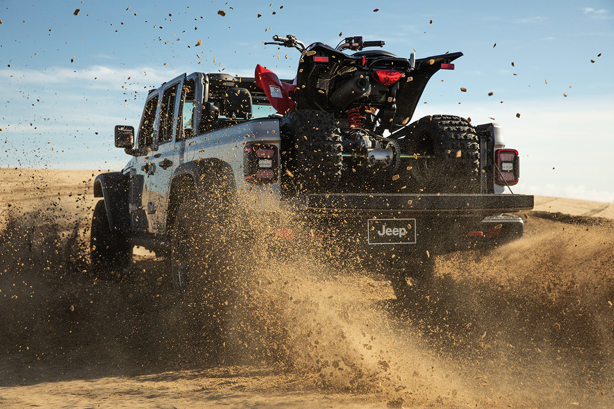 Rear shot of a silver Jeep Gladiator driving through a desert kicking up sand with an ATV in the pickup bed.