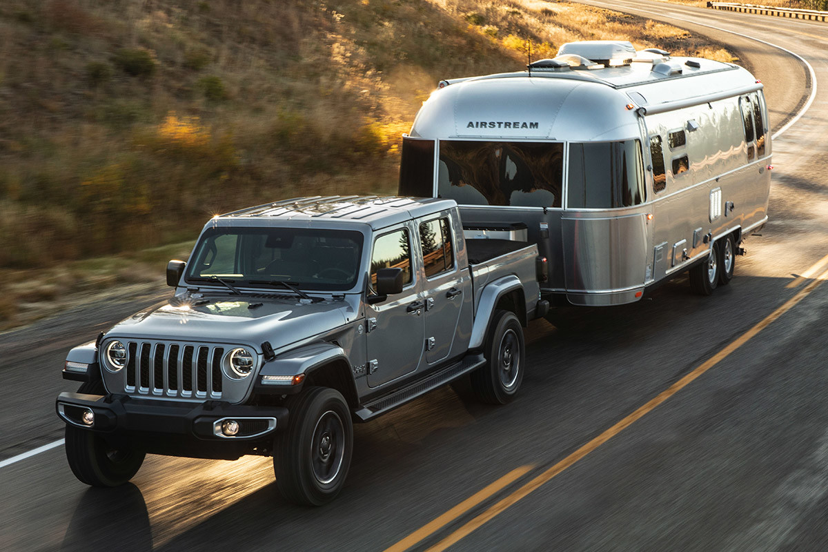Silver Jeep Gladiator towing a trailer while driving down a road alongside trees.