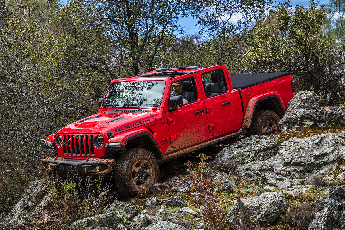 Red Jeep Gladiator driving down rocky off-road terrain with trees in the background.