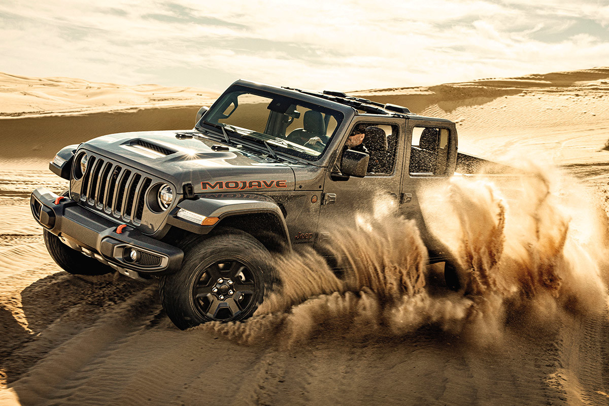 Silver Jeep Gladiator Mojave drives through a desert landscape on a sunny day, kicking up sand.