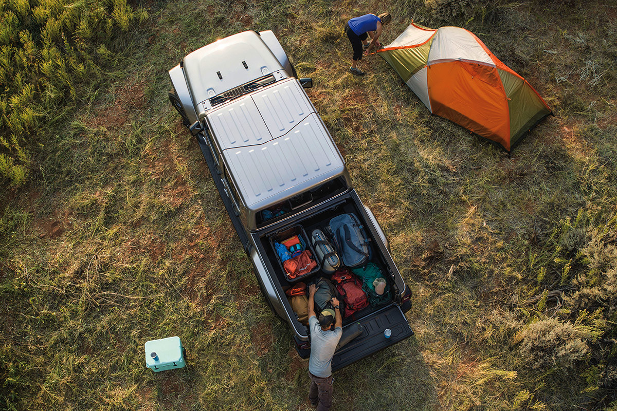 Jeep Gladiator parked on a grassy area. A man unpacks cargo from the pickup bed, while a woman sets up a camping tent next to the vehicle.