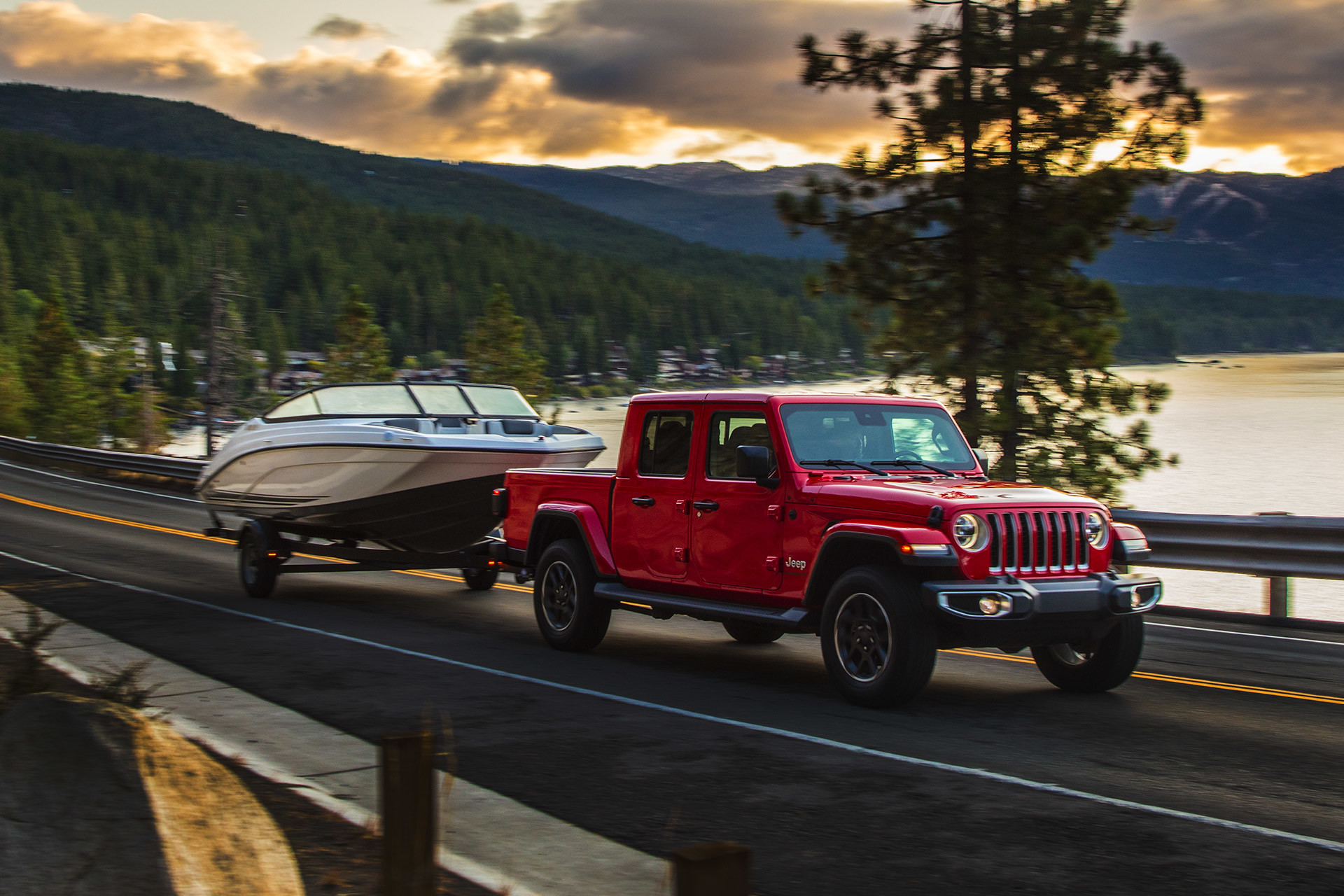 Red Jeep Gladiator towing a boat on a road with a lake and mountains in the background.