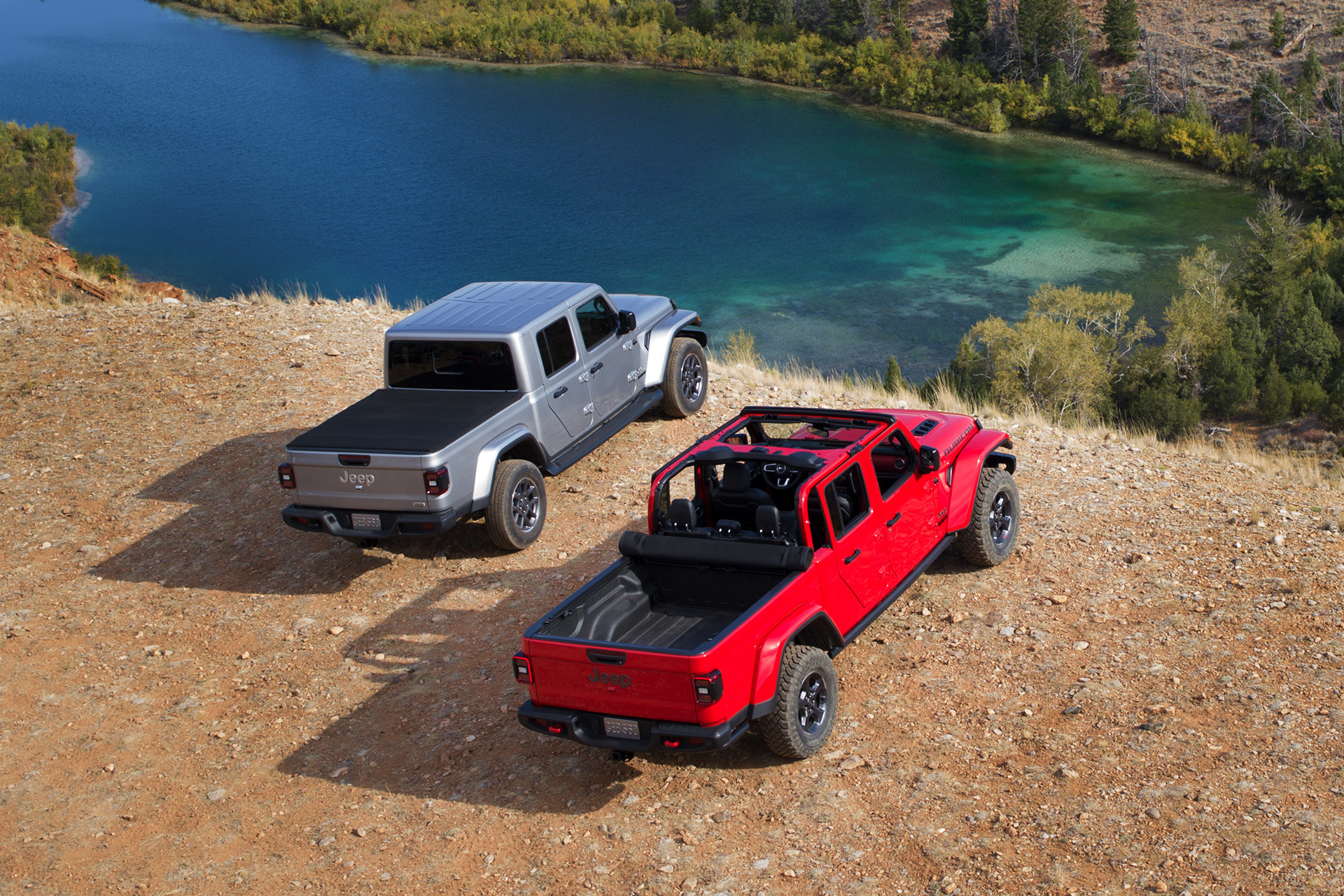 One silver Jeep Gladiator and one red Jeep Gladiator parked side by side overlooking a cliff, with the view of a small lake below.