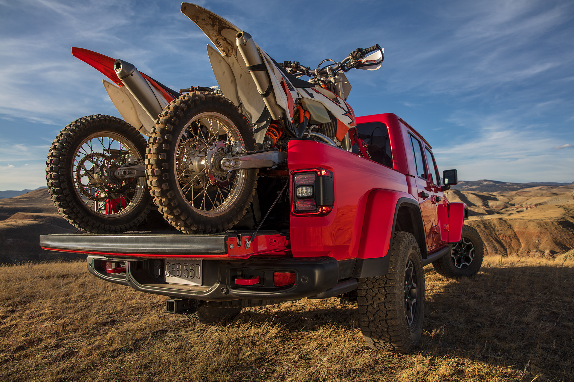 A 2022 Gladiator Rubicon in red parked on dirt patch with two dirt bikes in the truck bed.