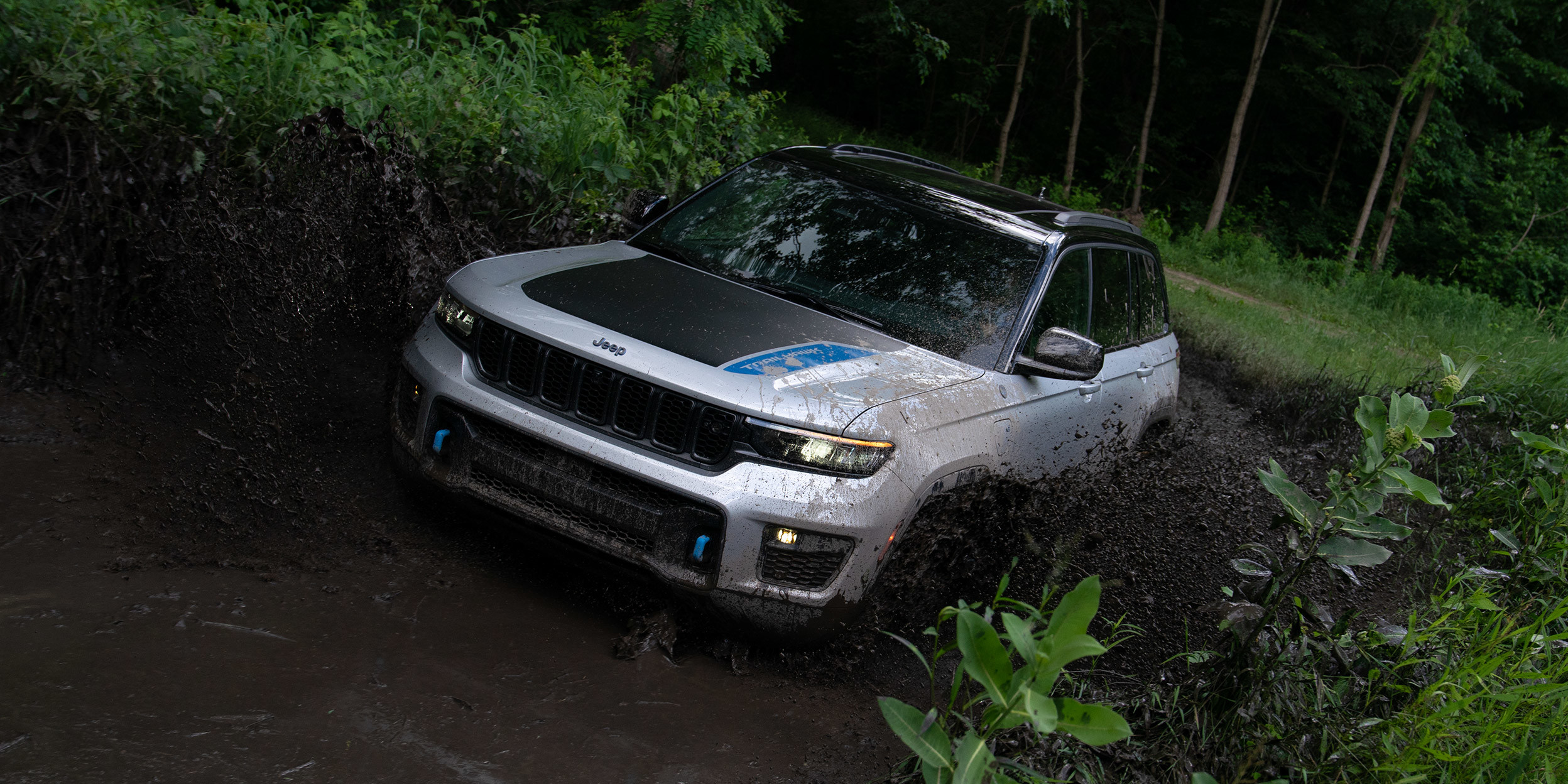 Un Jeep Grand Cherokee Trailhawk 4xe de 2022 de couleur argentée est conduit sur un chemin de terre à travers une forêt verte.