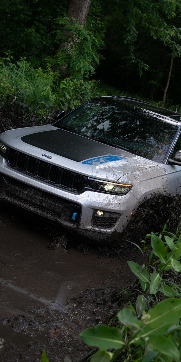 Un Jeep Grand Cherokee Trailhawk 4xe de 2022 de couleur argentée est conduit sur un chemin de terre à travers une forêt verte.