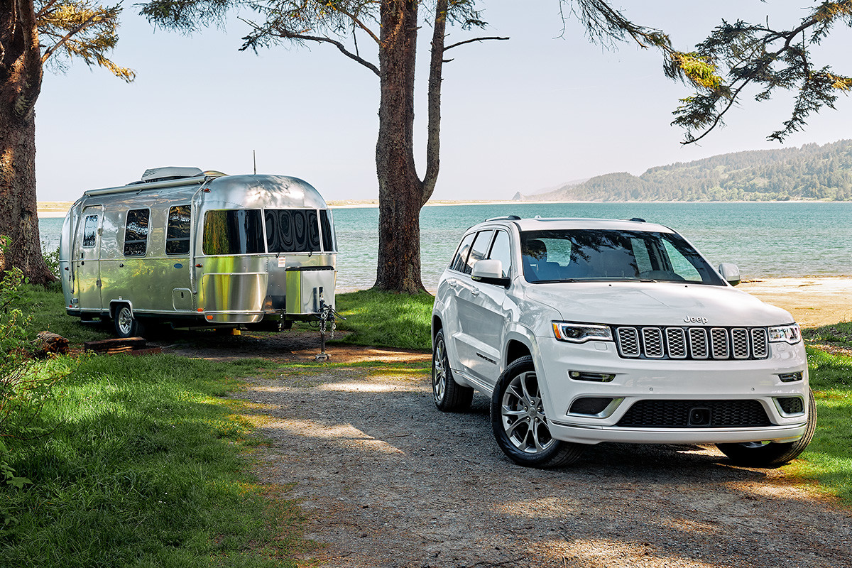 White 2020 Jeep Grand Cherokee, with a silver trailer attached at the back, parked on a gravel road with a beach coast in the background.