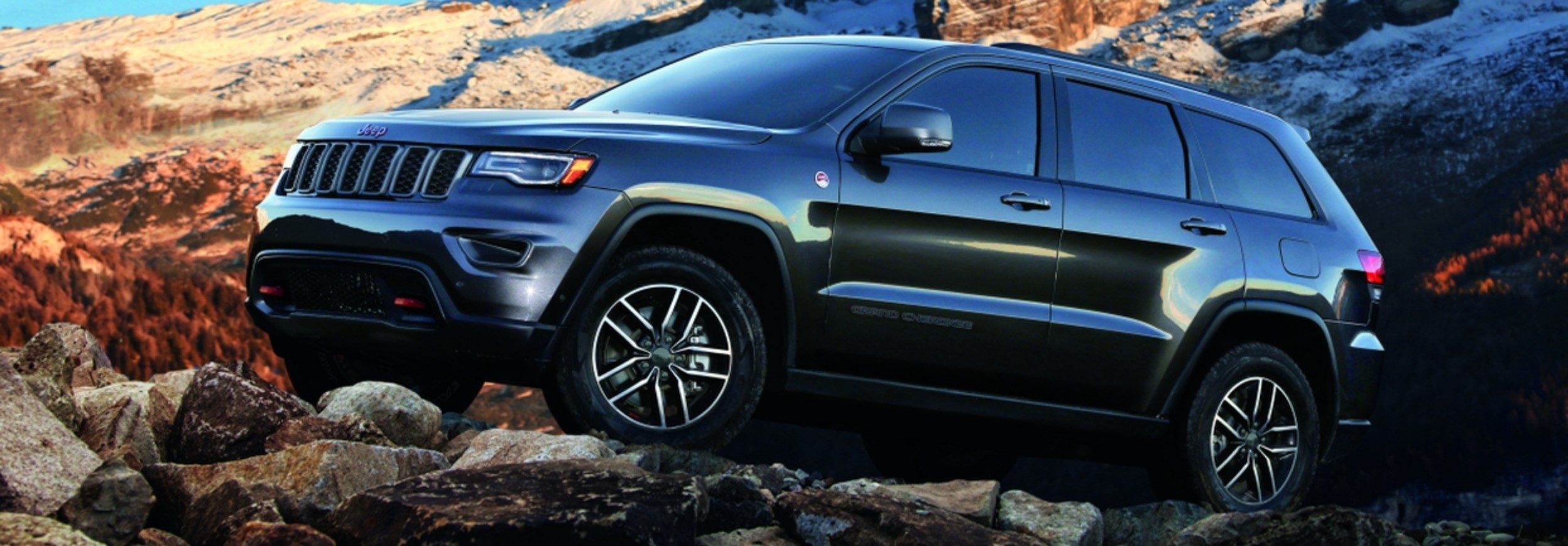 Metallic grey Jeep Grand Cherokee parked on a rocky surface with mountains in the background.
