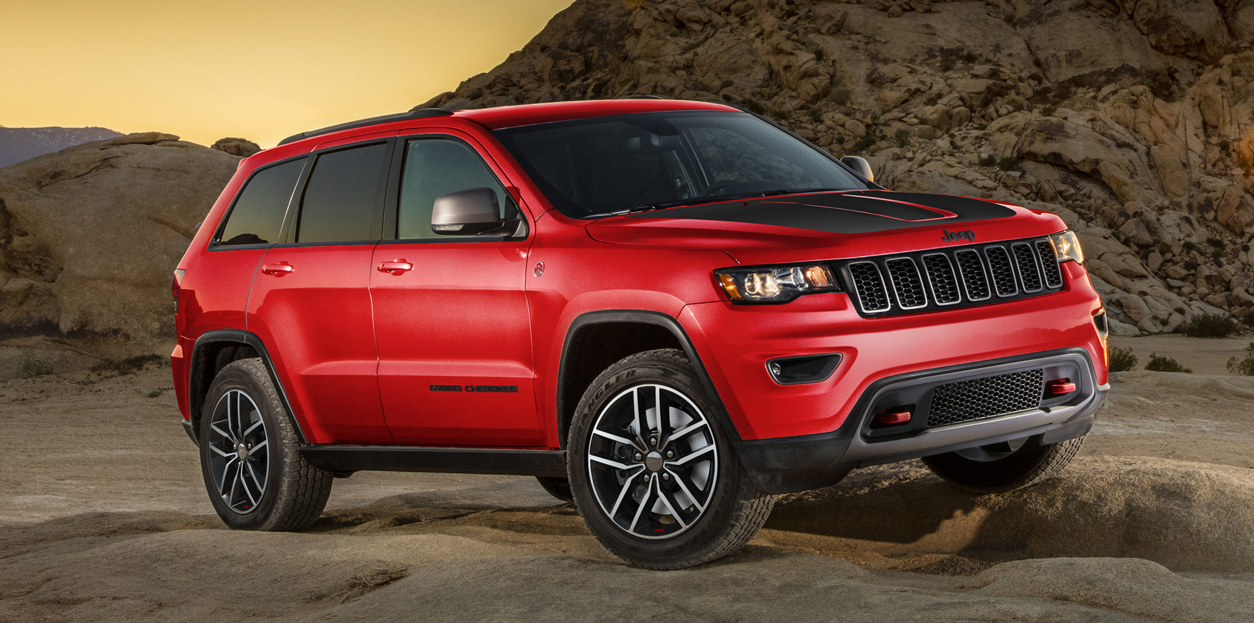 A red Jeep Grand Cherokee, parked on large boulders with mountains in the background.