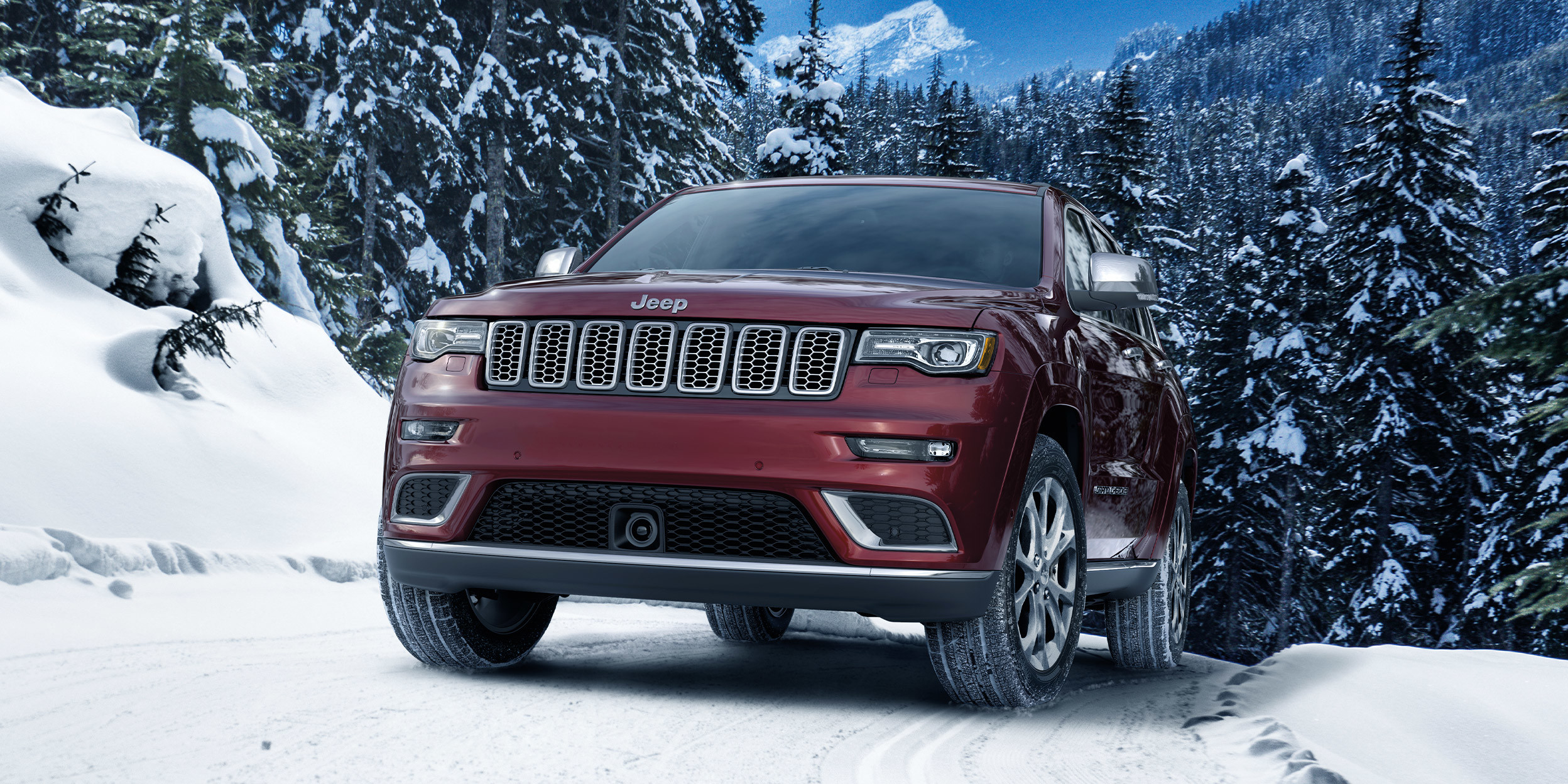 Grand Cherokee Summit rouge velours garé sur une route d’hiver enneigée, entouré d’arbres et de montagnes, le jour.