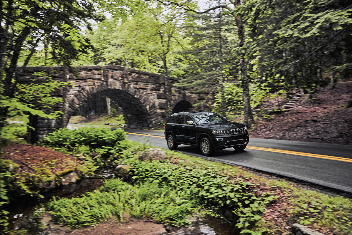 Grand Cherokee WK Limited 2022 roulant sur une route bordée de forêts, avec un pont de pierre derrière.