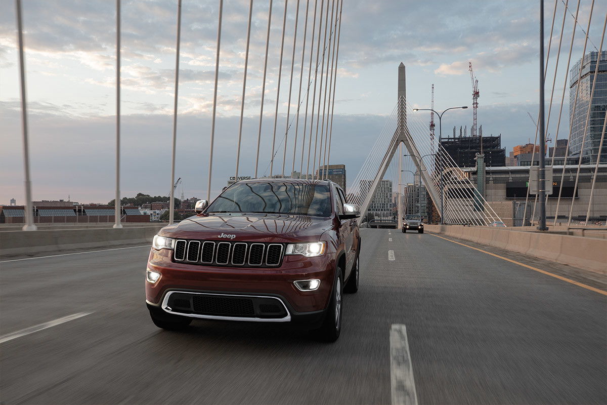 2022 Grand Cherokee Limited in Velvet Red Pearl driving on bridge with lights on, cityscape and cloudy sky in the distance.