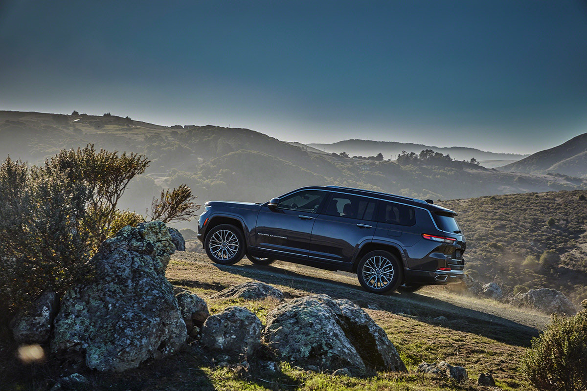 Vue latérale du tout nouveau Grand Cherokee L 2021 gris stationné sur un terrain verdoyant surplombant des vallées montagneuses voilées de brume.