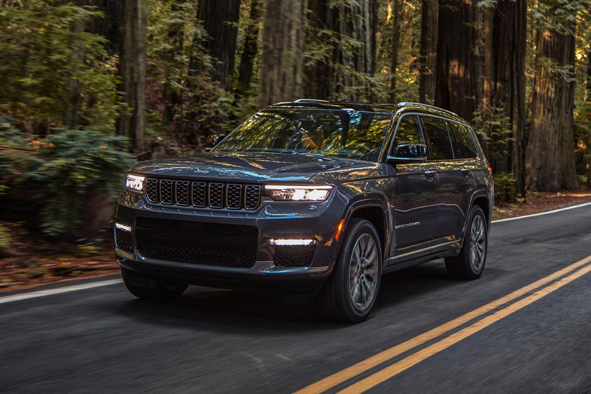 Vue de face d’un tout nouveau Grand Cherokee L 2021 gris roulant sur une route dans une forêt. Ultraminces, les phares à DEL brillent intensément.