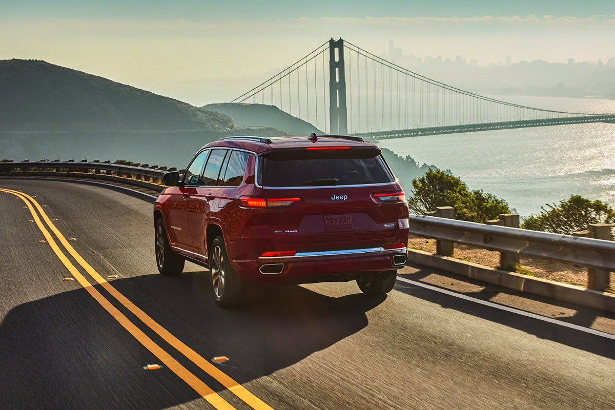 Vue arrière du tout nouveau Grand Cherokee L 2021 rouge roulant sur une route surplombant l’océan. Un pont suspendu apparaît au loin.