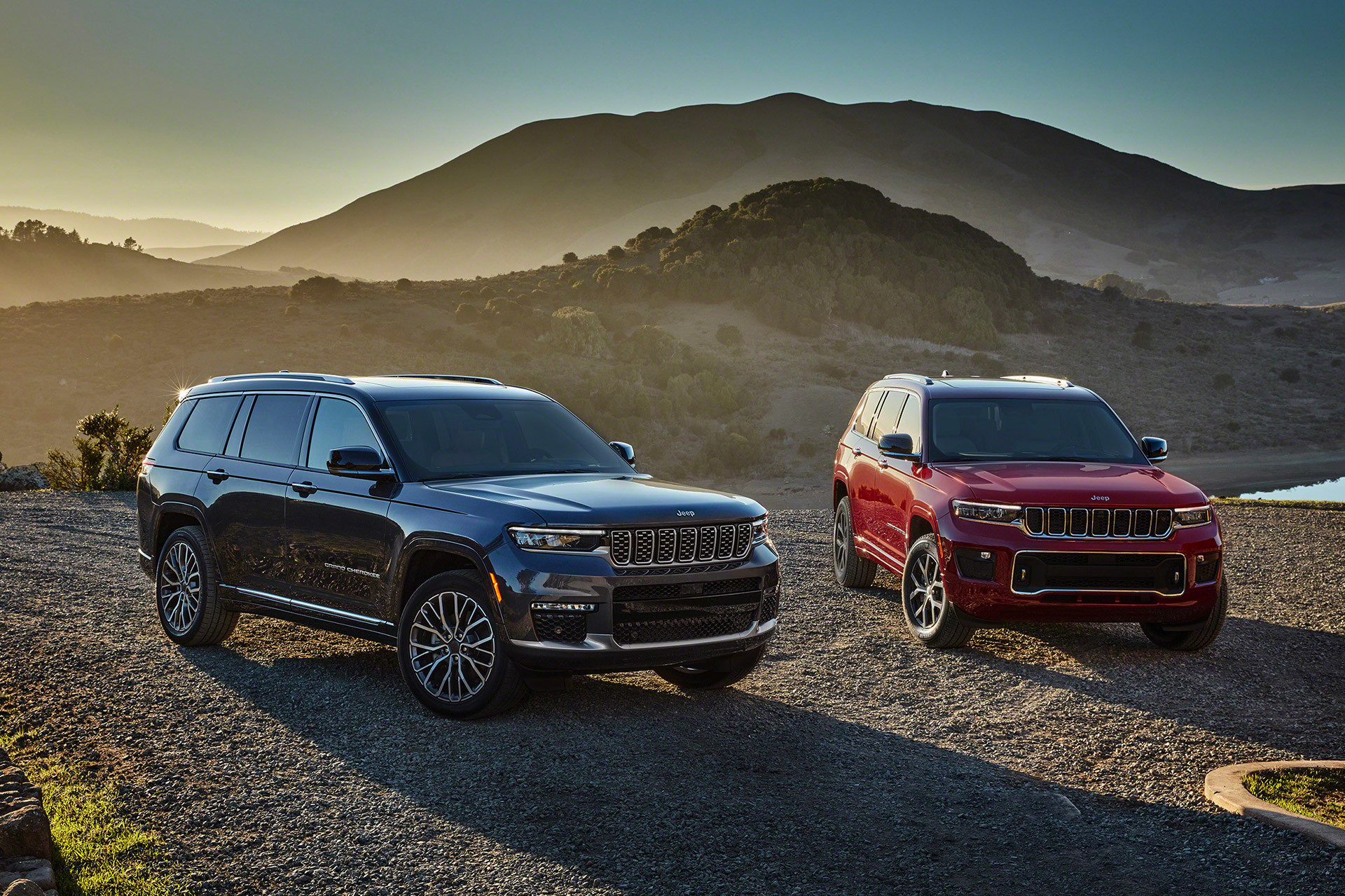 Vue de deux tout nouveaux Grand Cherokee L 2021 sur une route de gravier sur fond de montagnes. Un véhicule est gris et l’autre rouge.