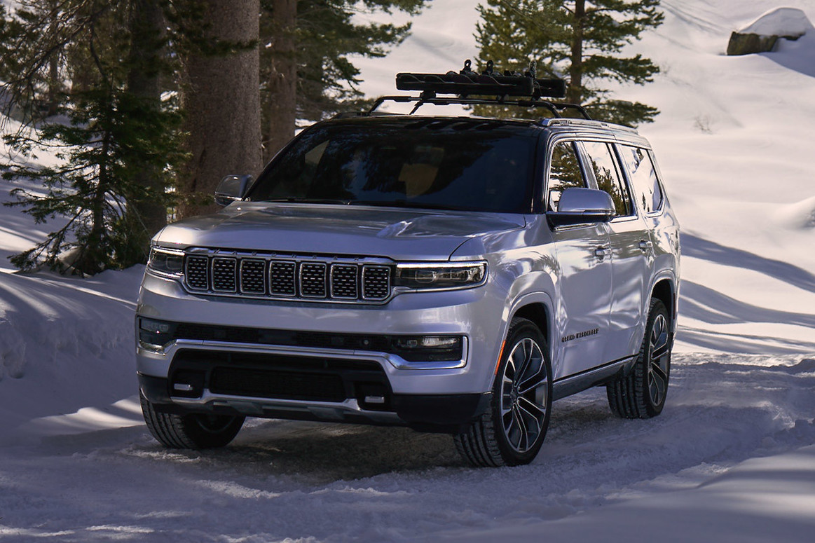 Un tout nouveau Grand Wagoneer 2022 blanc, garé au pied d’une colline enneigée avec des arbres en arrière-plan.