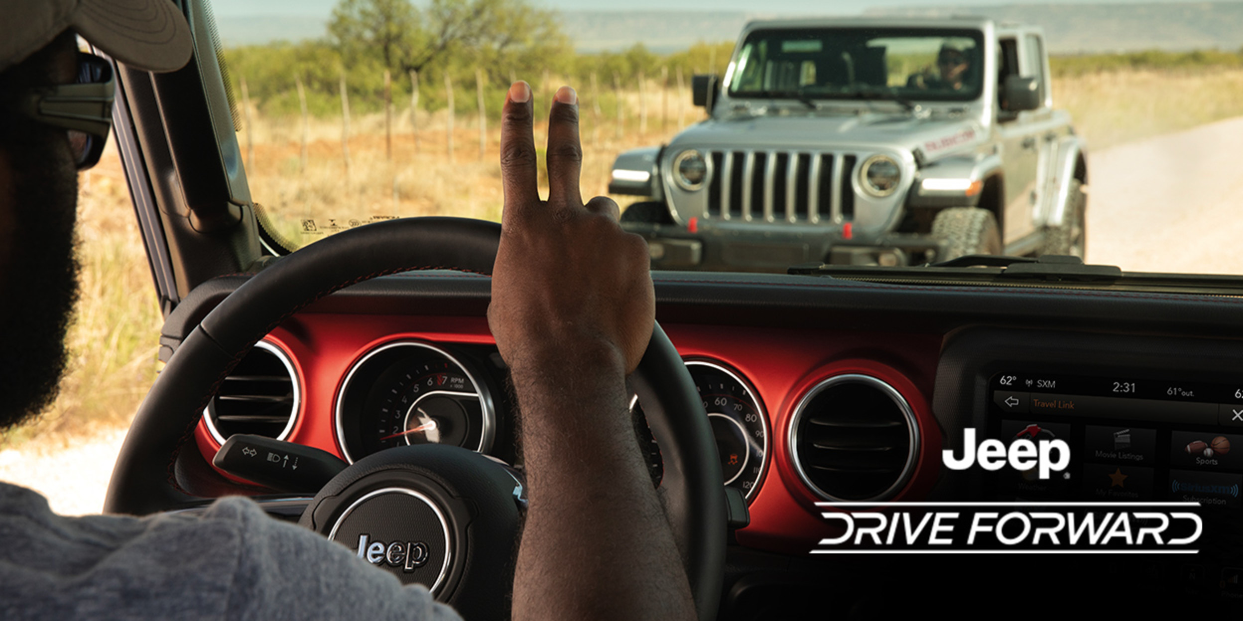 A driver in a Jeep Wrangler driving down a dirt road, greeting another driver driving a grey Jeep Wrangler on the other side of the road.