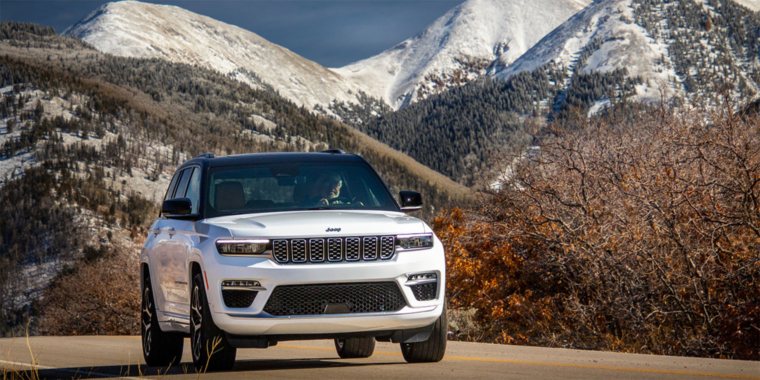 Front view of a white 2023 Jeep Grand Cherokee driving on a paved road with snow covered mountains in the background.
