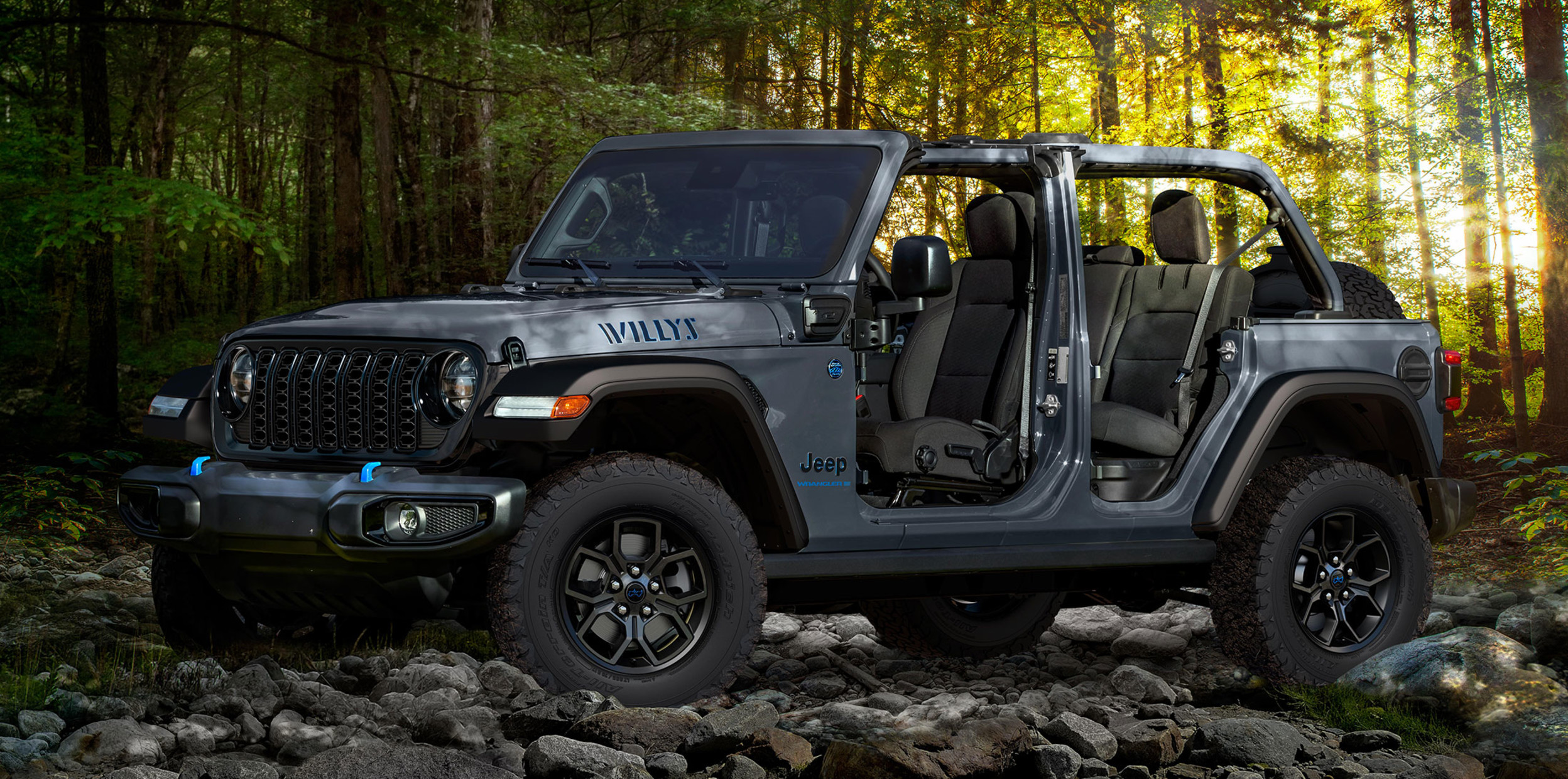 A profile view of a grey 2024 Jeep Wrangler parked on top of rocks inside a forest with trees and sunlight peeking through the trees in the background.