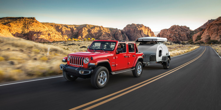 Profile view of a red 2023 Jeep Gladiator towing a closed trailer on a paved roadway with desert terrain shown behind.