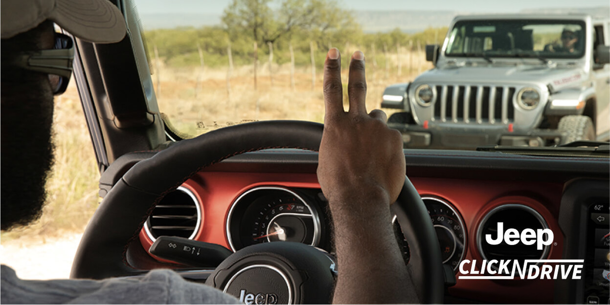 Jeep Click ’n Drive. A driver in a Jeep Wrangler driving down a dirt road, greeting another driver driving a grey Jeep Wrangler on the other side of the road.
